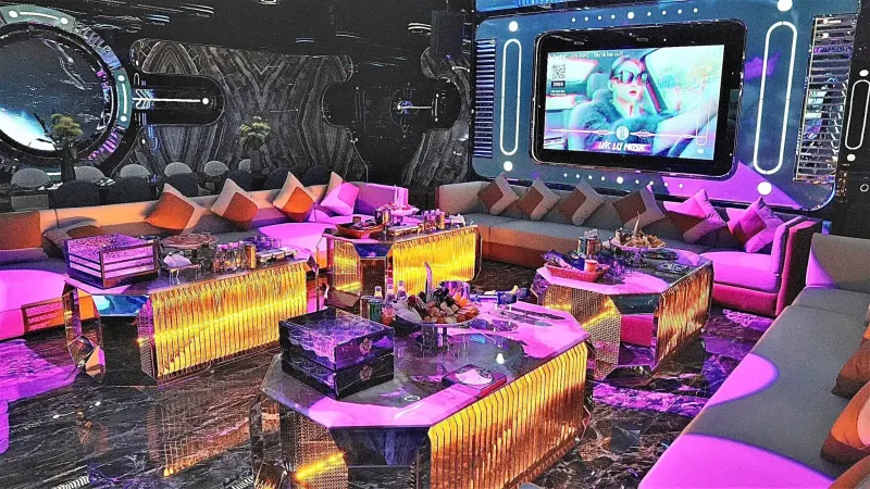 Phòng hát dưới ánh đèn tím hồng, sofa gối tựa, bàn đèn vàng và màn hình lớn